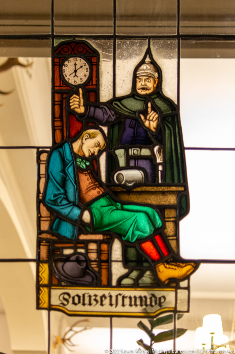 Stained-glass tavern scene with a sleeping seated man, standing figure, wall clock, and large drinking vessel.