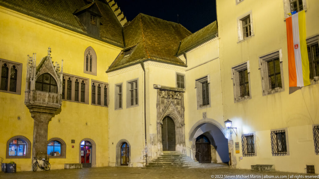 Night courtyard of Regensburg's Altes Rathaus with ornate Gothic portal, projecting stone oriel, pointed windows, and civic flags.