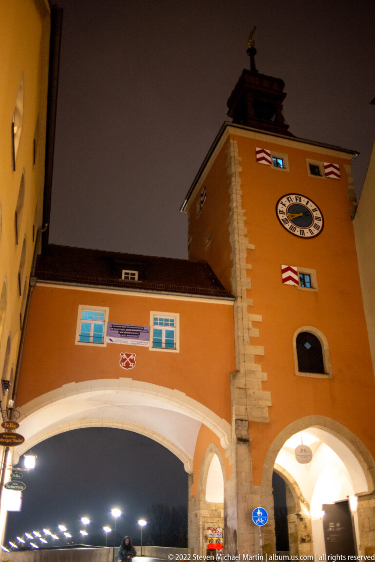 Illuminated orange Brückturm at night with clock, red-and-white shutters, stone corner blocks, and broad gateway arch.