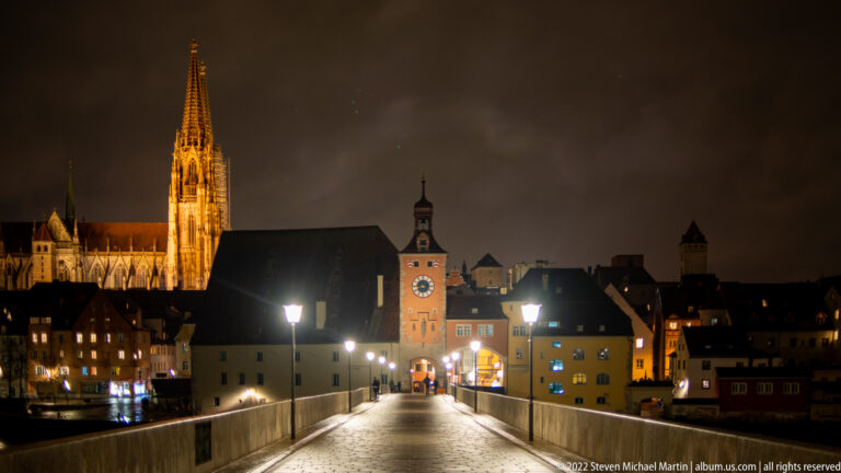 Night view along Regensburg's Stone Bridge toward the illuminated Brückturm, with St. Peter's Cathedral glowing above the skyline.