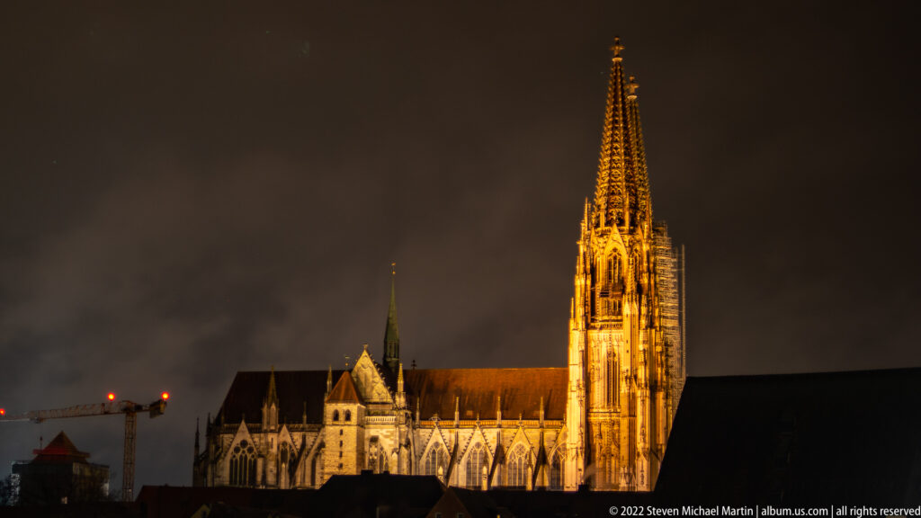 Illuminated Gothic spire and roofline of St. Peter's Cathedral against a cloudy night sky in Regensburg.