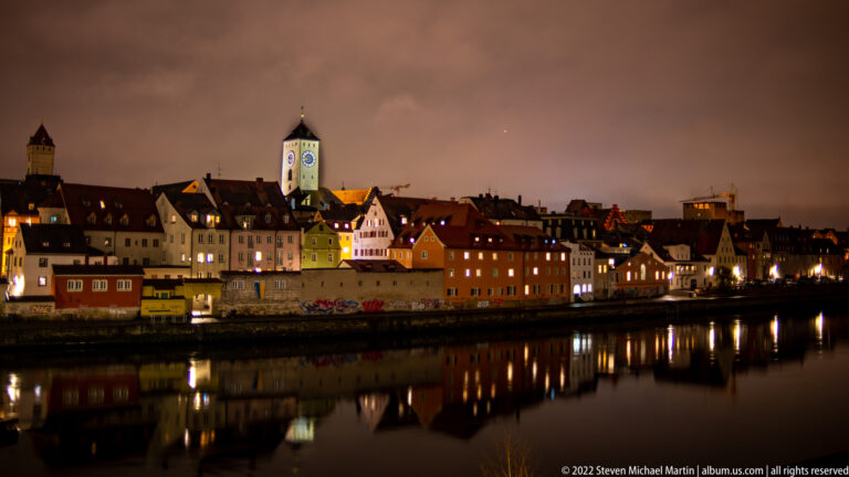 Colorful Regensburg riverfront buildings and square clock tower reflected in the Danube at night.
