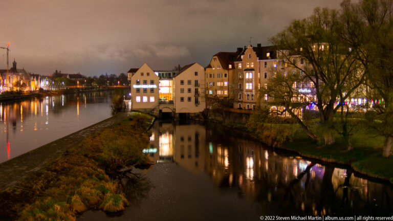 Historic riverside houses reflected in a narrow Danube channel at night, with trees and grassy banks.