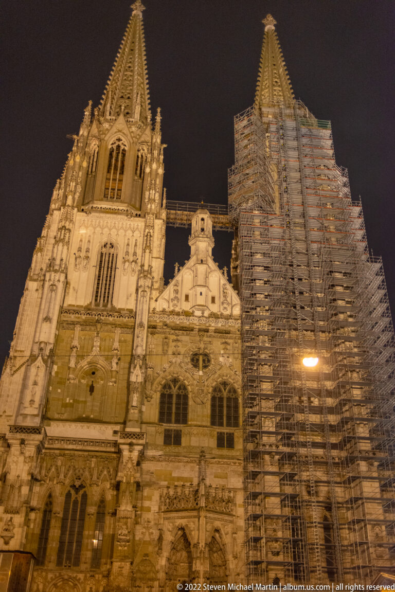 Illuminated Gothic west front of Regensburg Cathedral with one tower wrapped in extensive scaffolding.