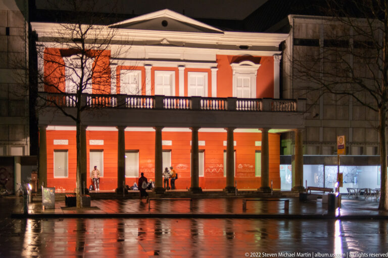 Red-and-white Neoclassical building with pediment, colonnade, and balcony reflected in wet pavement at night.