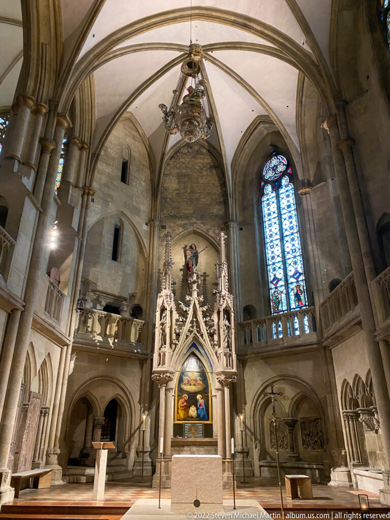 Church sanctuary with rounded arches, ribbed vaults, galleries, tall stained glass, and an ornate central Gothic shrine.