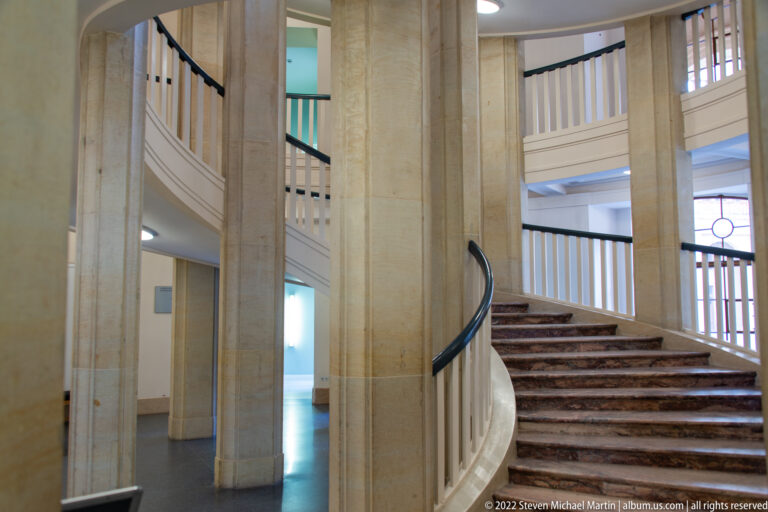 Curving stone staircase with pale columns, dark steps, black handrail, and white balustrades.