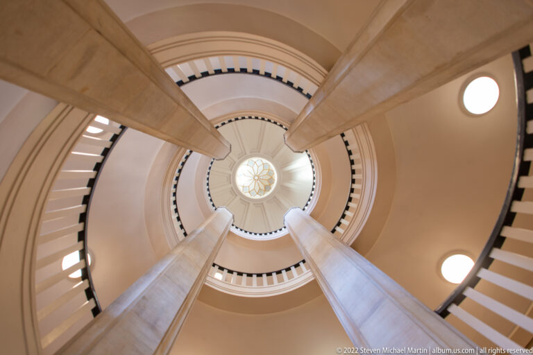 Upward view through a spiral staircase toward a circular glazed opening, with pale supports and round lights.