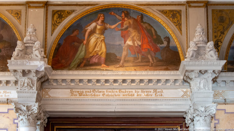 Arched mural of two historical figures above a doorway, with German inscription and carved ornament.