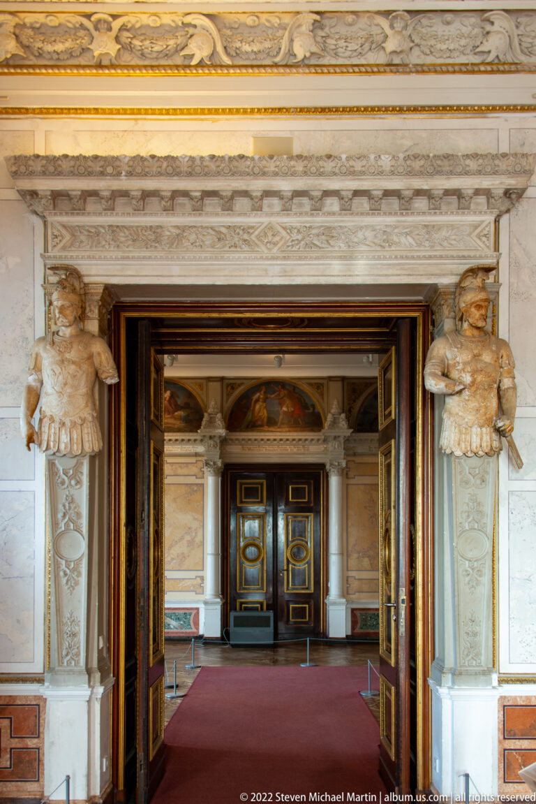 Ornate marble doorway with carved figures opening into a red-carpeted sequence of palace rooms.