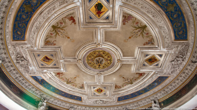 Ornate circular ceiling with white plasterwork, painted floral panels, gilding, and central medallion.