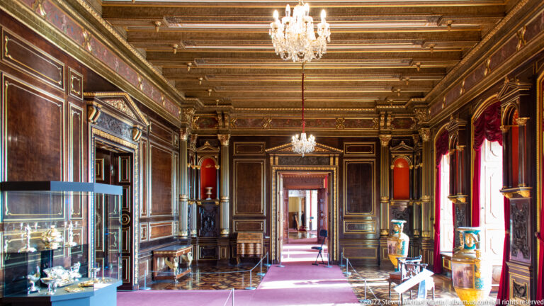 Long dark wood-paneled palace room with chandeliers, red curtains, display cases, and an aligned doorway.