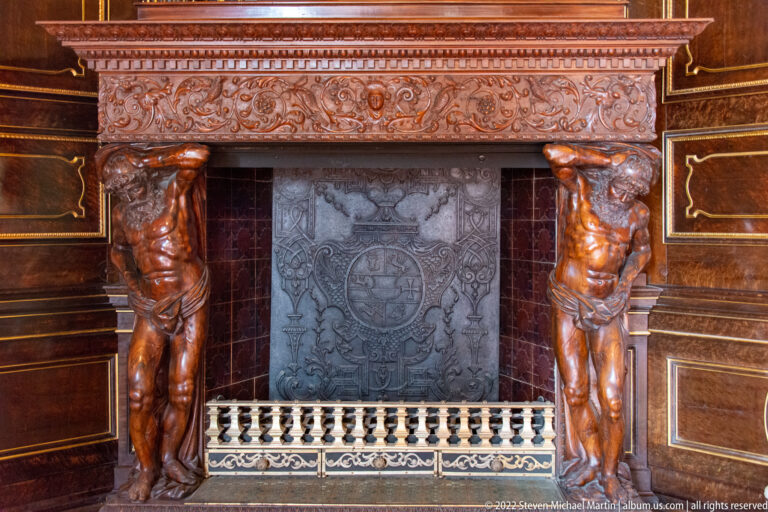 Ornate dark fireplace with two carved male supports, heraldic fireback, and pale grate.