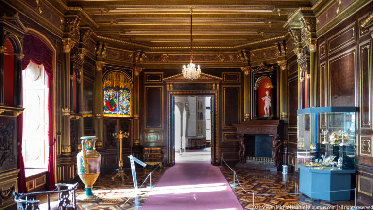 Dark paneled palace room with stained glass, carved fireplace, chandelier, vitrines, and sunlit parquet floor.