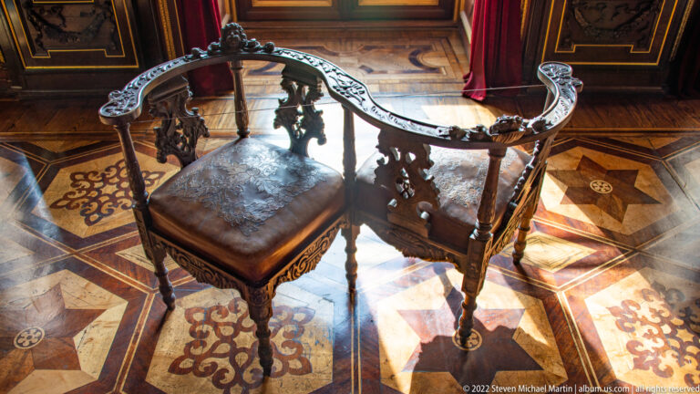 Ornately carved dark wooden armchair with leather seat on an inlaid parquet floor.