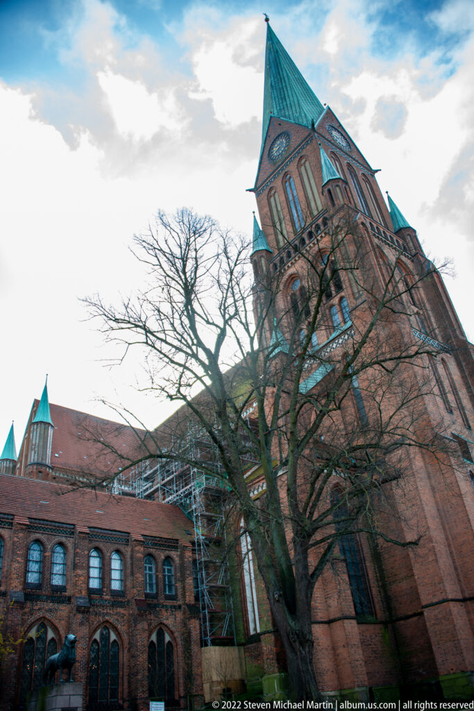 Low-angle view of Schwerin Cathedral’s brick tower, green roof, bare tree, and adjoining nave.