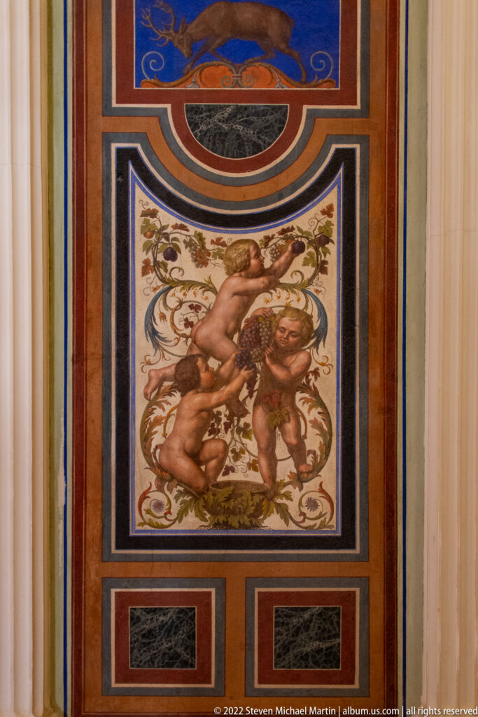 Square painted panel of two putti amid foliage, framed by multicolored ornamental borders.