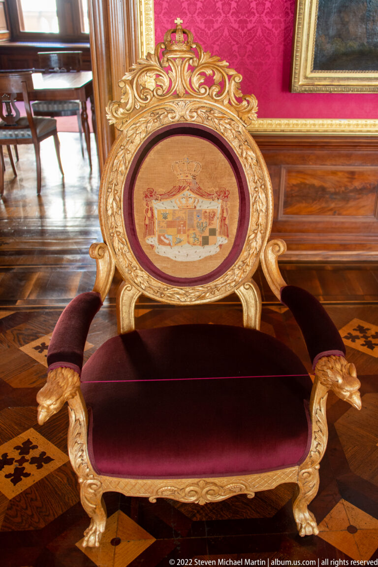 Gilded ceremonial armchair with red velvet upholstery and painted heraldic shield on the oval back.