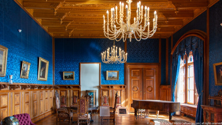 Blue state room with carved wood paneling, chandeliers, framed paintings, chairs, and tall windows.