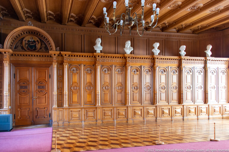 Wood-paneled Ancestral Gallery with portrait busts, chandelier, carved doors, and geometric parquet floor.