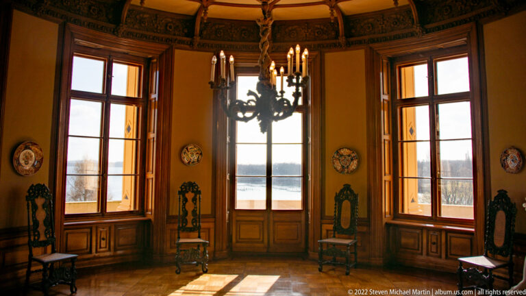 Curved palace room with three tall windows, carved high-backed chairs, wall plaques, and dark chandelier.