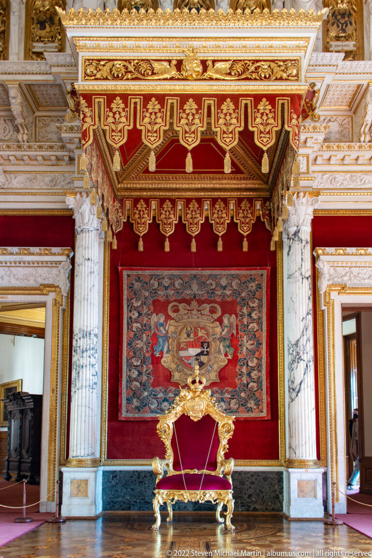 Gilded throne beneath red-and-gold canopy, heraldic textile, and paired marble columns.