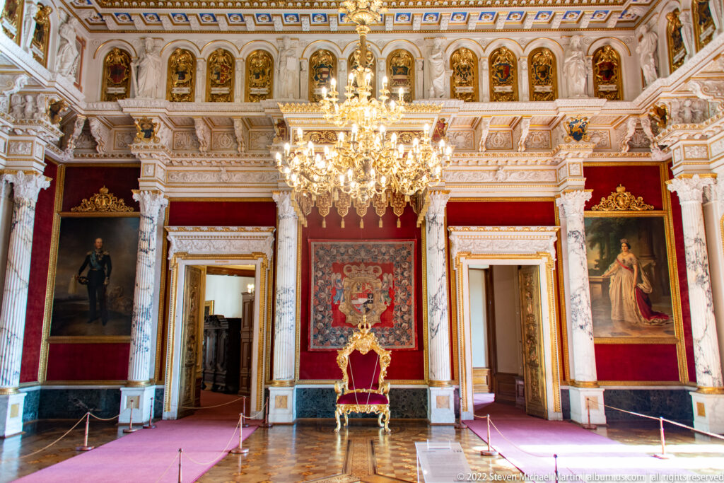Wide Throne Room view with central throne, chandelier, marble columns, portraits, red walls, and gilded decoration.