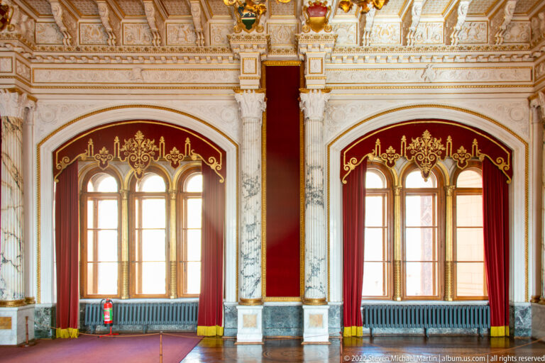 Two arched windows with red curtains, marble columns, gilded ornament, and polished floor in the Throne Room.
