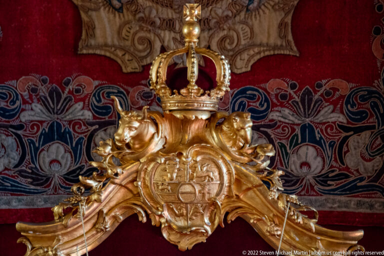 Close view of gilded crowned coat of arms and sculpted supporters on the throne against red and blue textile.