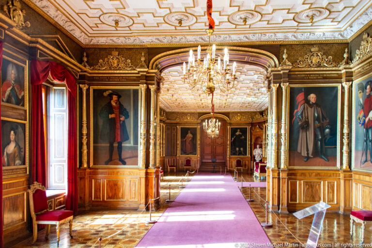 Long Ancestral Gallery with full-length portraits, chandeliers, gilded frames, red curtains, and purple runner.