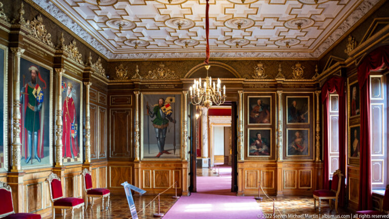 Portrait-lined Ancestral Gallery with chandelier, paneled walls, doorways, and red chairs.