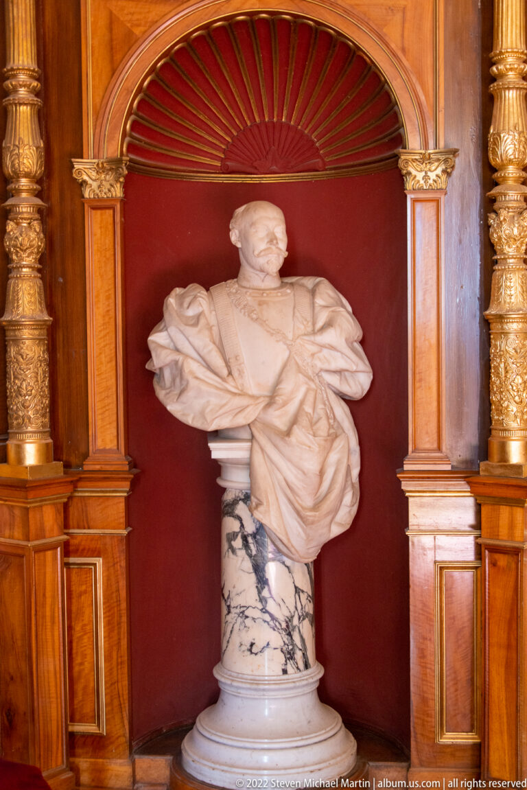 White marble male portrait bust in a red niche with gilded columns and colored marble pedestal.