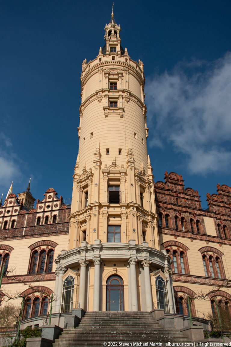 Low-angle view of Schwerin Castle’s tall cylindrical tower above red-brick gables and pale arcades.