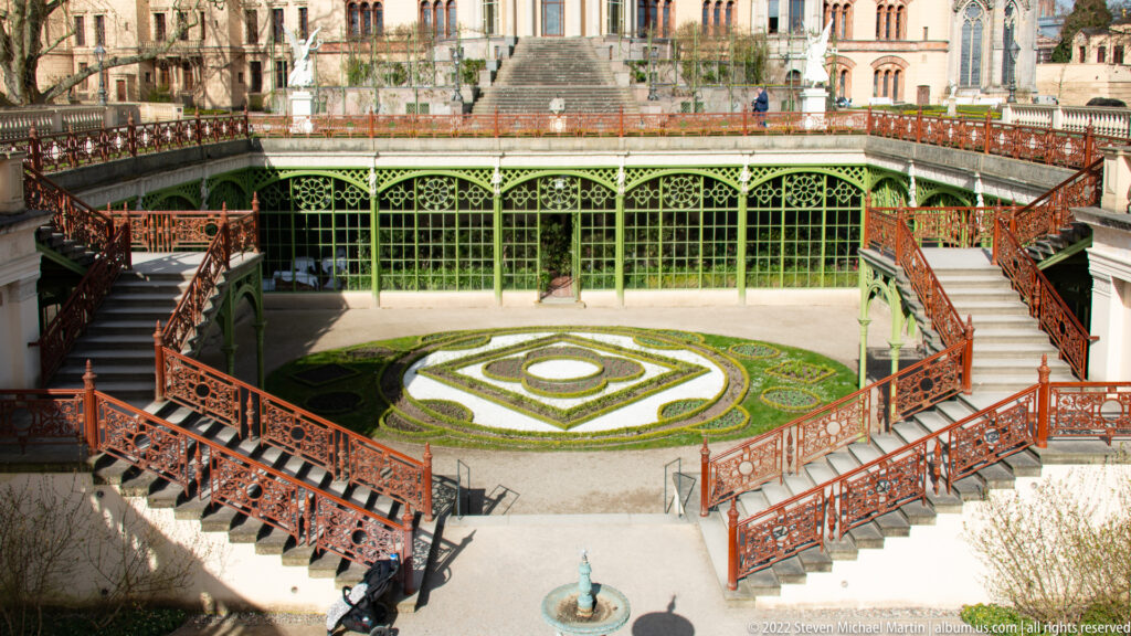 Formal Orangerie courtyard with geometric parterre, green iron glazing, paired red staircases, and small fountain.