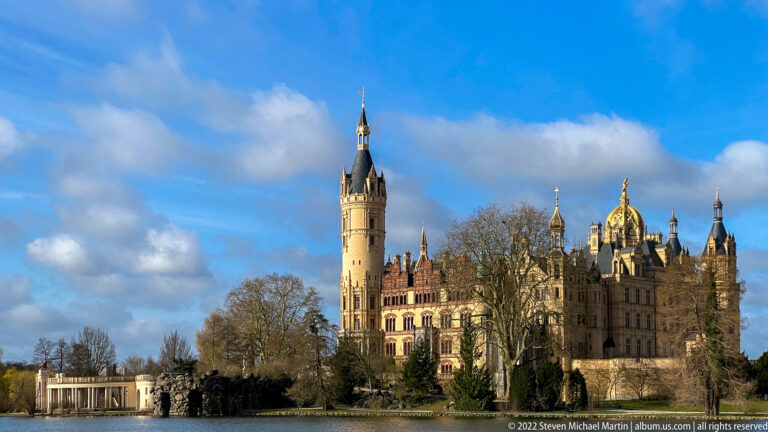 Schwerin Castle across Burgsee with tall tower, turrets, gilded domes, bare trees, and blue sky.