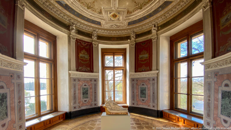 Round palace room with tall windows, red decorative panels, ornate ceiling, and reclining white sculpture on a pedestal.