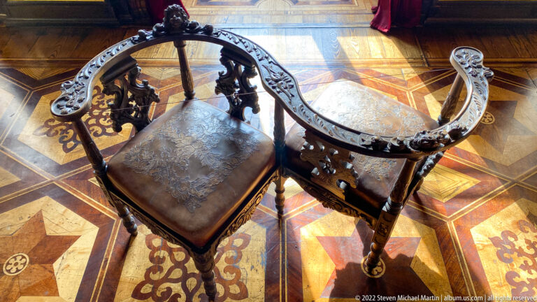 Dark carved corner armchair with curved pierced back and leather seat standing on richly patterned parquet.