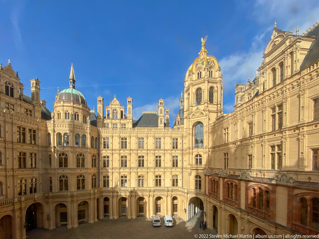 Sunlit inner courtyard of Schwerin Castle enclosed by ornate pale façades, arcades, towers, and varied rooflines.