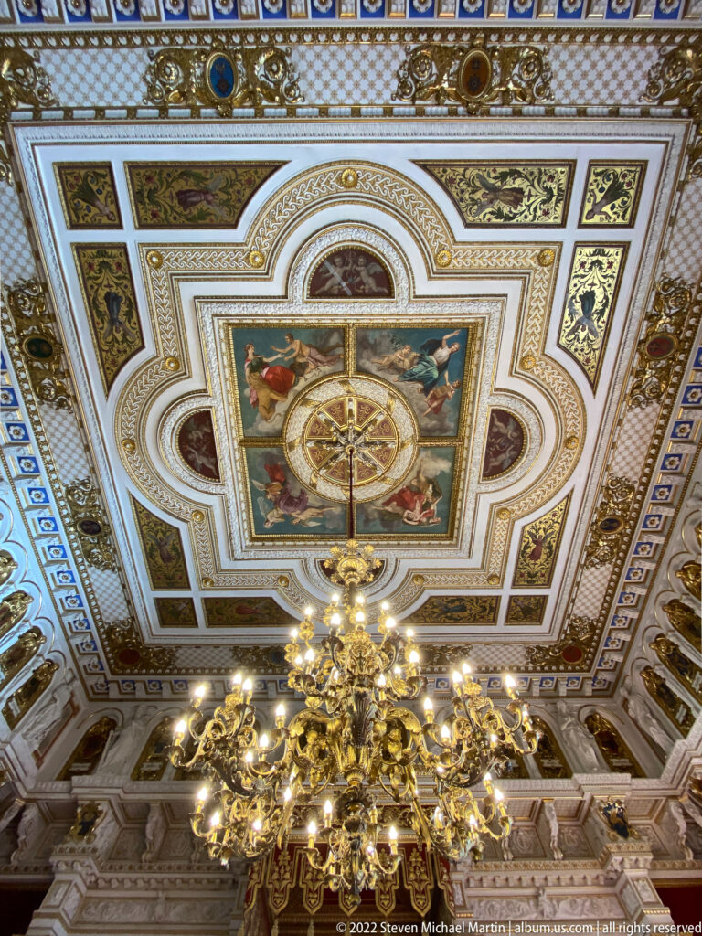 Ornate coffered ceiling with gilded moldings, painted figural panels, central circular design, and large gilt chandelier.