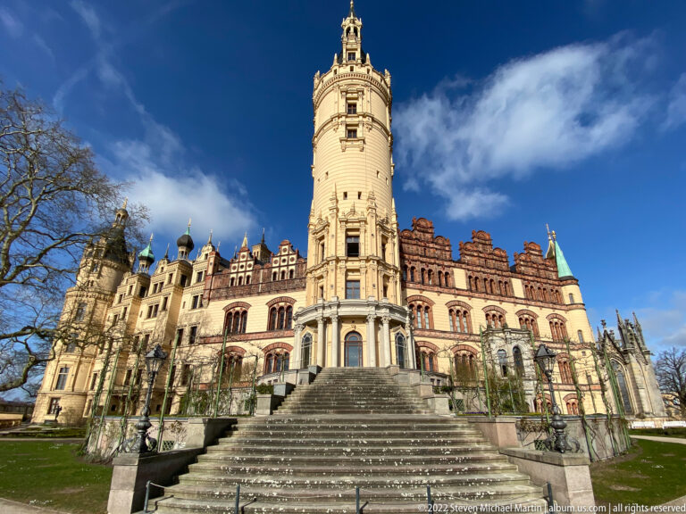 Broad staircase rising toward Schwerin Castle’s ornate garden façade and tall cylindrical main tower beneath blue sky.
