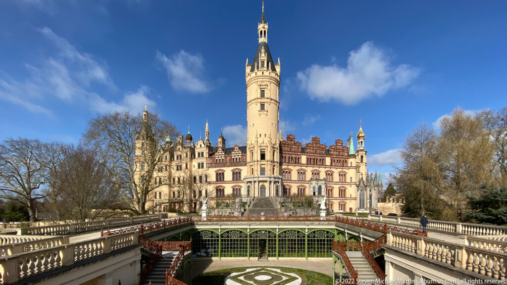Schwerin Castle centered beyond balustraded terraces and the sunken Orangerie garden, with its tall tower against blue sky.