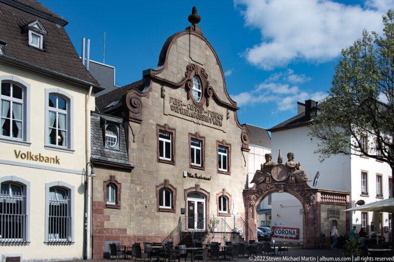 Curving historic stone façade beside a sculpted gateway, with café seating and neighboring buildings under a blue sky.