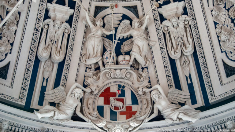 White stucco figures and drapery surrounding a red-and-white heraldic shield against dark blue panels.