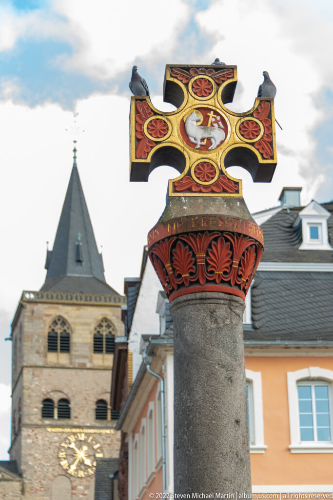 Painted cross on a stone column in Trier's Hauptmarkt, with a Romanesque church tower and rooftops behind.