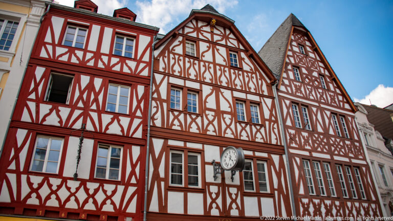 Three steep-gabled half-timbered houses with red timber framing, pale infill panels, and a projecting clock.