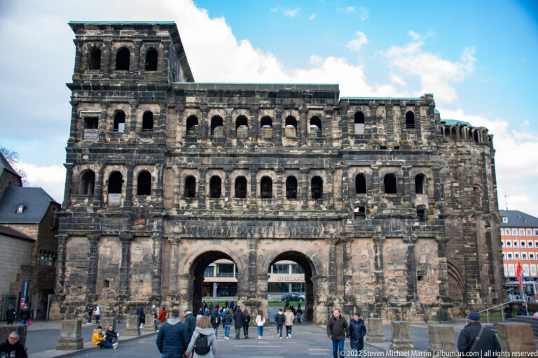 Front view of Trier's Porta Nigra with massive dark stone walls, rows of arches, twin gateways, and visitors below.