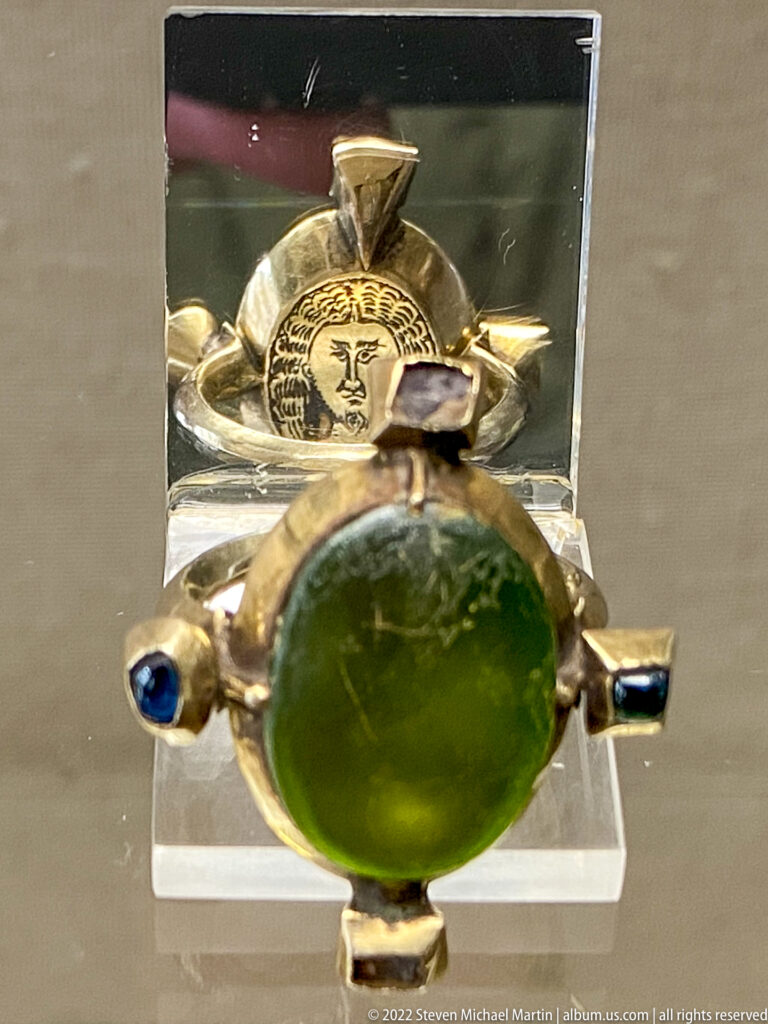 Gold cross-like devotional object with a large oval green cabochon, blue stones, and a circular face medallion behind.