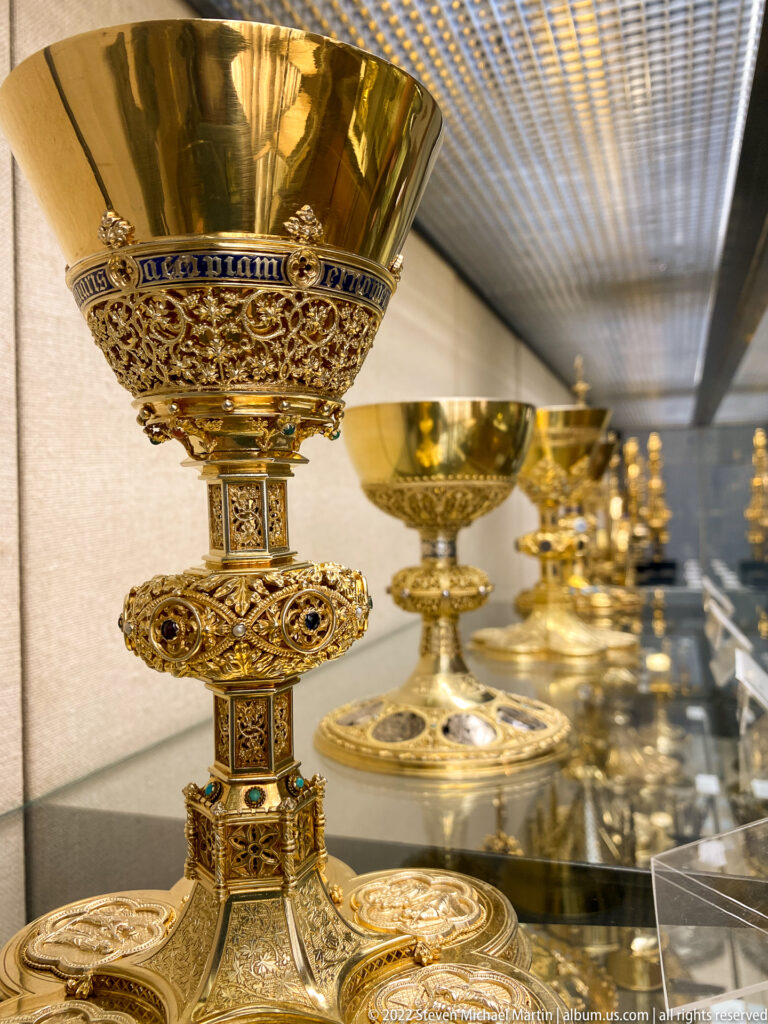 Gold liturgical chalice with filigree, colored stones, decorated stem, and additional chalices visible in the case behind.
