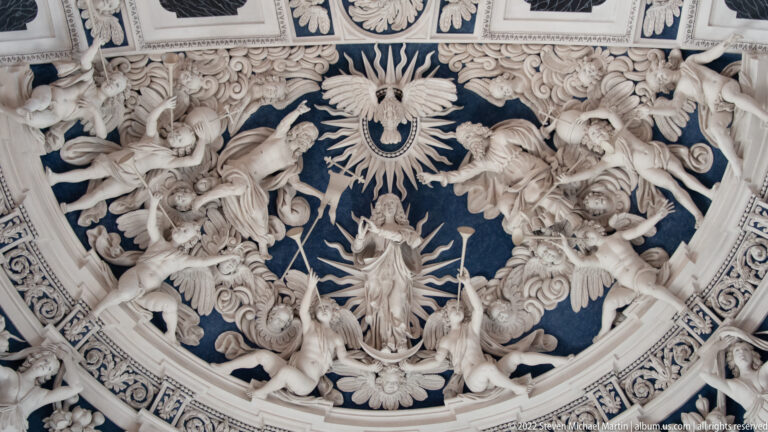 Radiant central sacred figure surrounded by angels, clouds, foliage, and white stucco ornament on a dark blue background.
