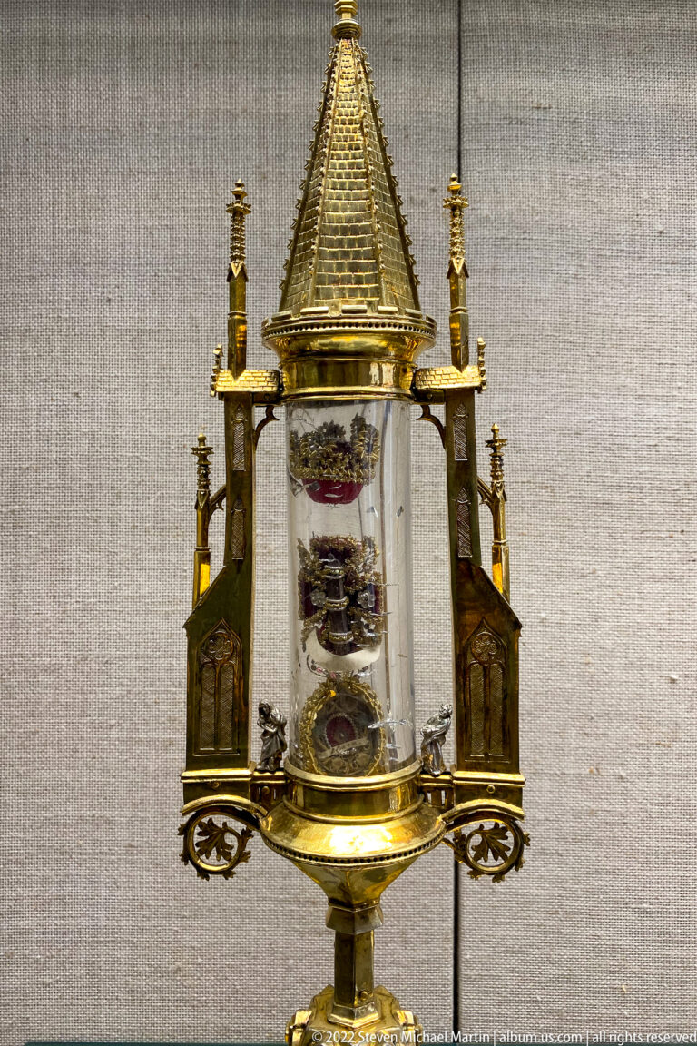 Small gilded architectural reliquary with pointed roof, corner turrets, tracery, and relics visible through a clear central chamber.